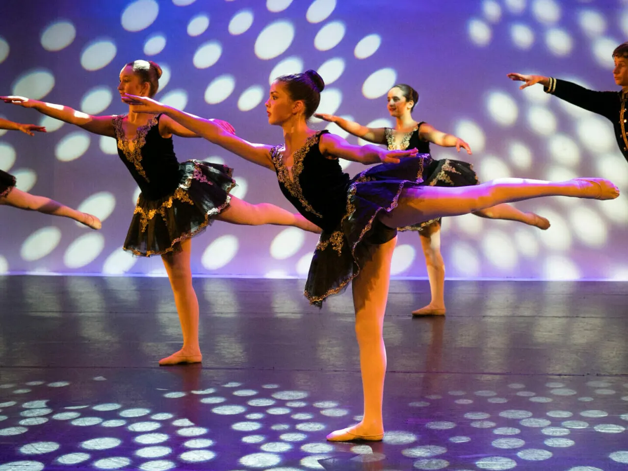 Young ballet dancers performing in synchronised formation on stage, wearing black and gold costumes against purple lighting with circular projections.