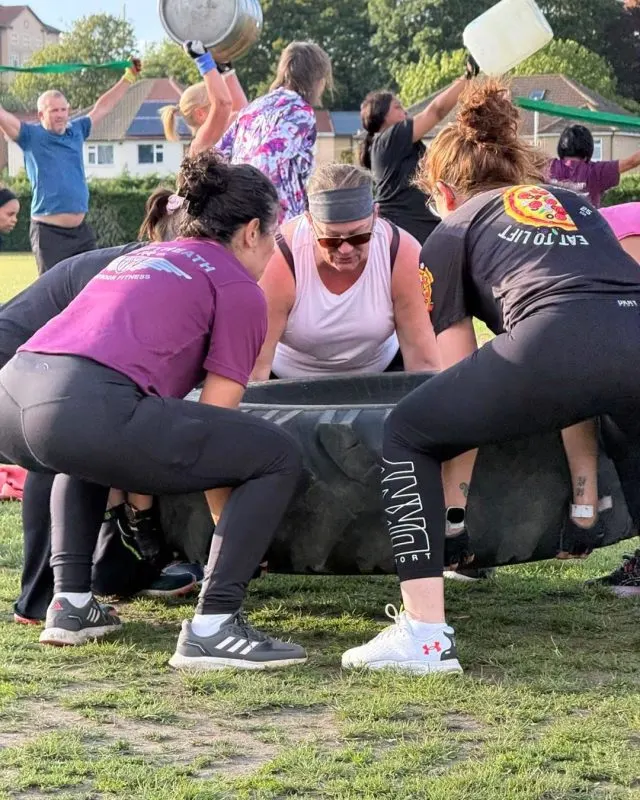 Group of people doing outdoor fitness training, lifting a large tyre together in a garden setting.