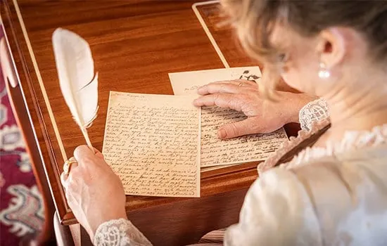 Woman in period costume holding a quill and reading handwritten manuscript at wooden desk.