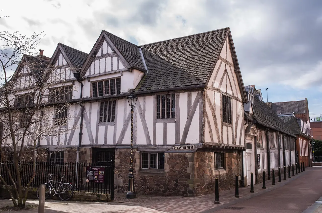 Half-timbered medieval building with white and dark timber framing, stone base, and slate roof under cloudy sky.
