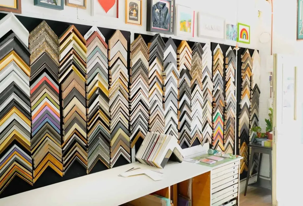 Display of picture frame moulding samples in various colours and finishes arranged on a black wall behind a counter.