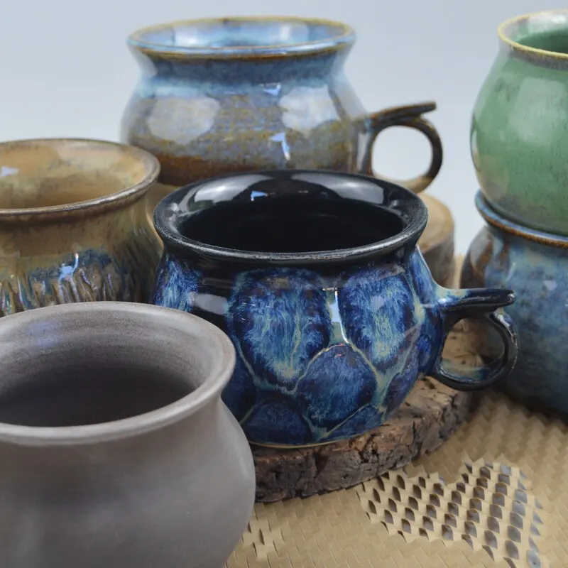 Collection of glazed ceramic vessels in blue, green and neutral tones displayed on a woven surface.