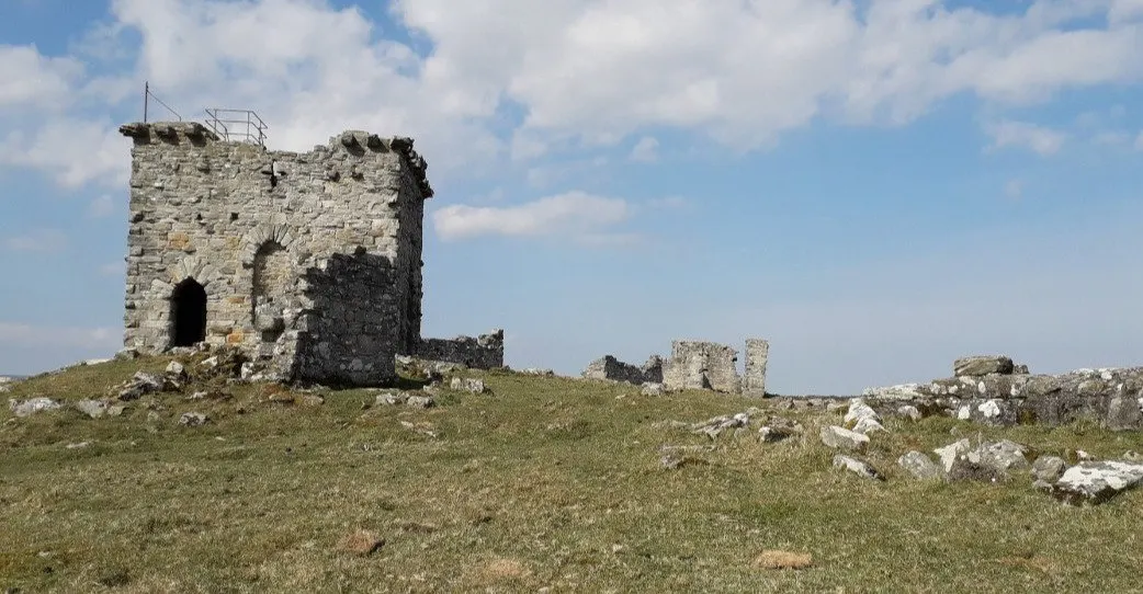 Ruined stone tower and fortifications on a grassy hillside dotted with rocks under blue sky.