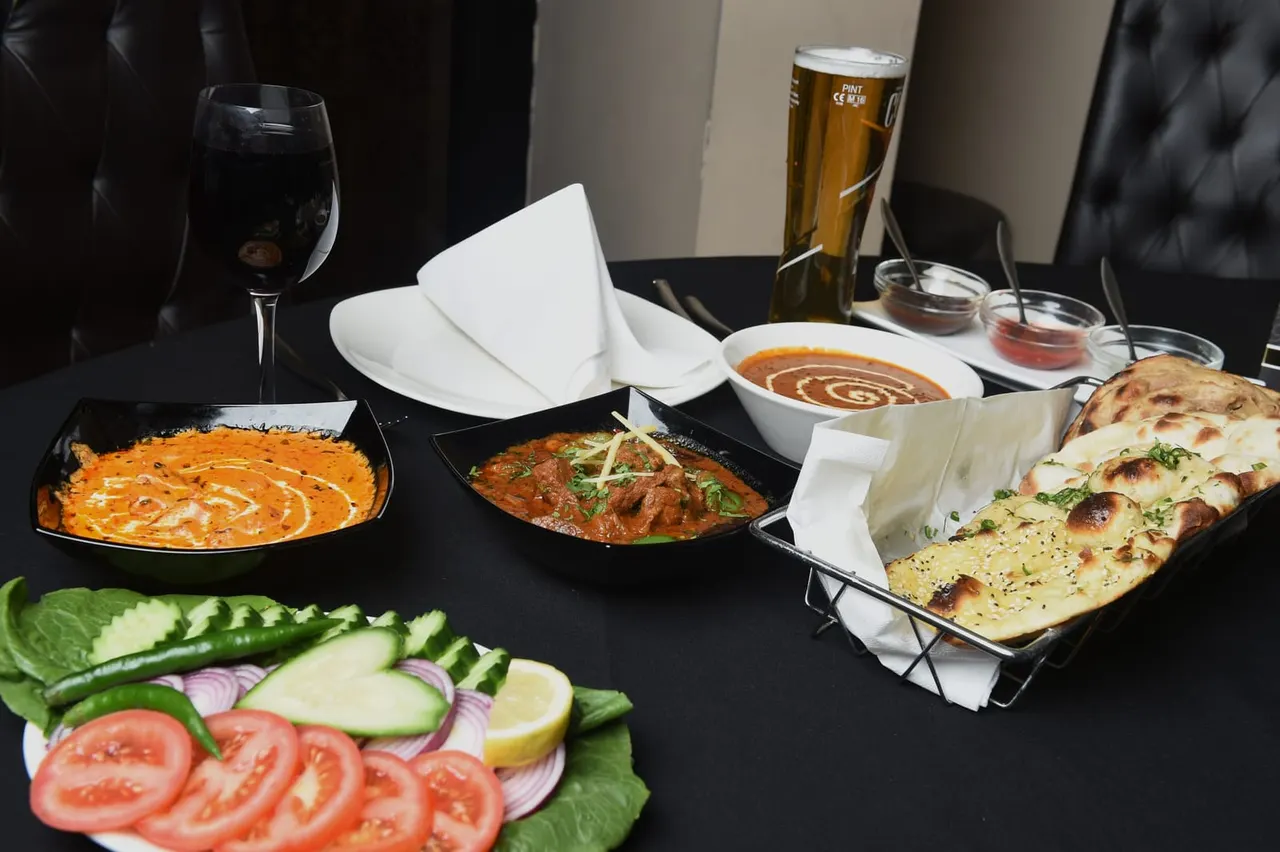 A table laid out with Indian dishes including curry, meat curry, naan bread, fresh salad, and accompaniments, with wine and beer.