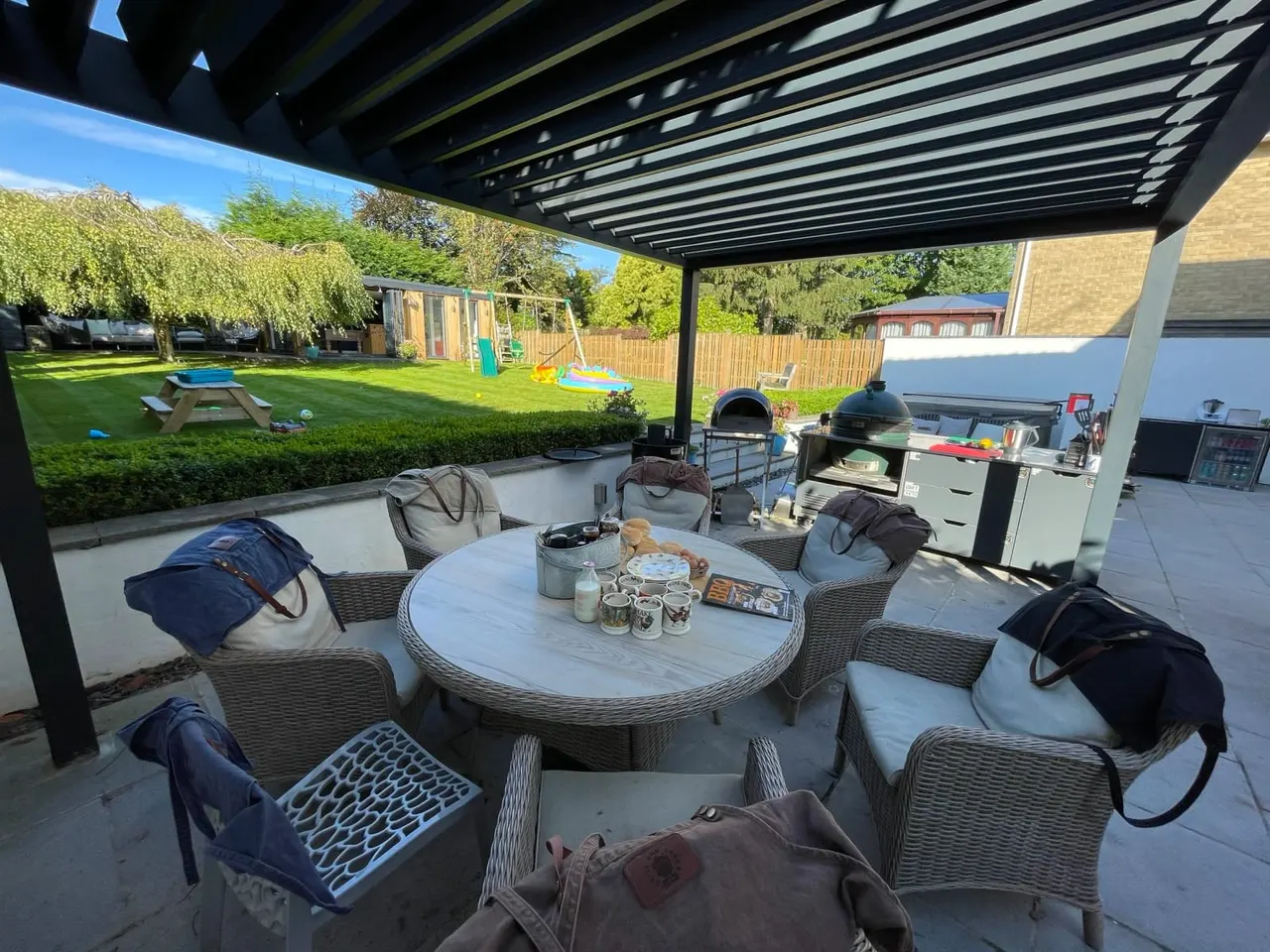 Outdoor patio area with rattan seating around a round table, pizza oven and barbecue, overlooking a family garden with play equipment and lawn.