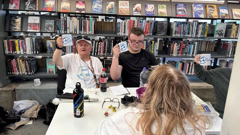 Two men sit at a table holding up badges whilst speaking to a woman with long hair, with shelves of DVDs and books visible behind them.