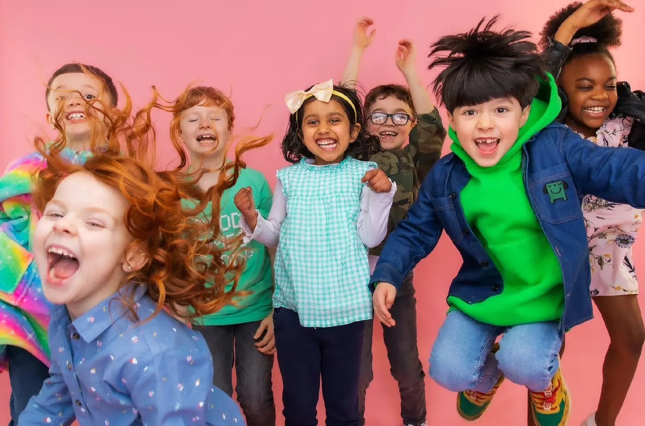 A group of children jumping and smiling joyfully against a bright pink background, celebrating together.