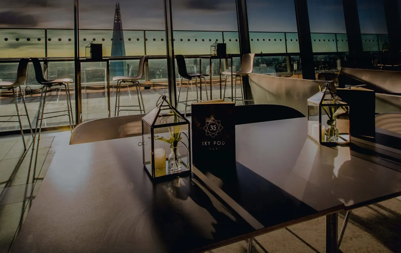 High-altitude bar seating area with floor-to-ceiling windows overlooking a city skyline and distant church spire.