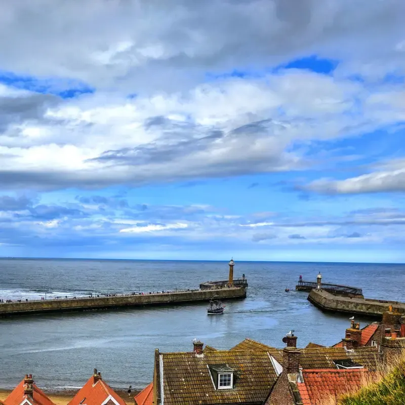Coastal harbour with stone piers, lighthouses and a small boat, viewed from clifftop cottages with red-tiled roofs.