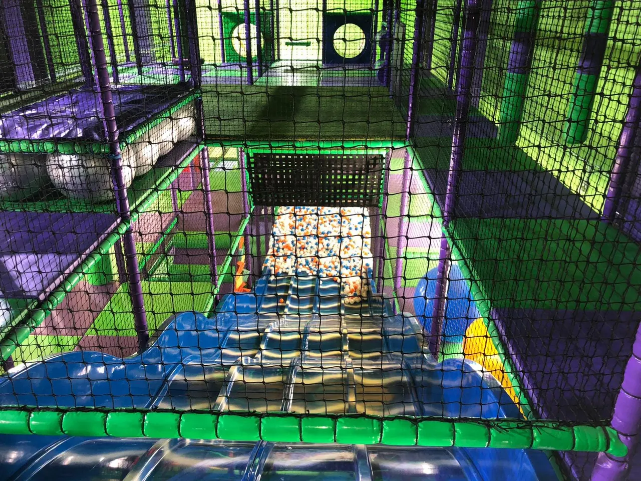 Interior view of a colourful soft play structure with netting, foam blocks and climbing elements lit in green and purple.