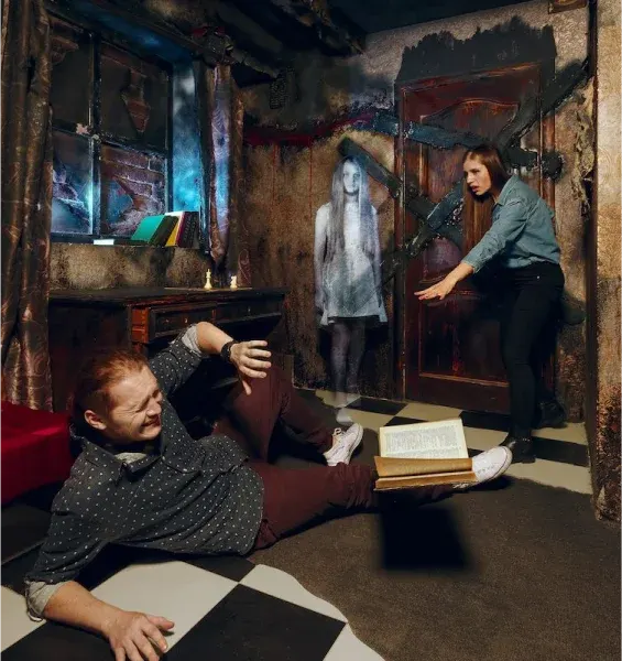 Two visitors interact with a ghostly figure projection in a dimly lit haunted room escape game environment.