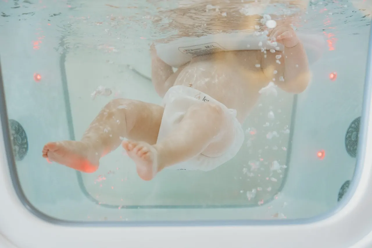 Baby submerged in warm water bath with bubbles and flower petals during spa treatment.