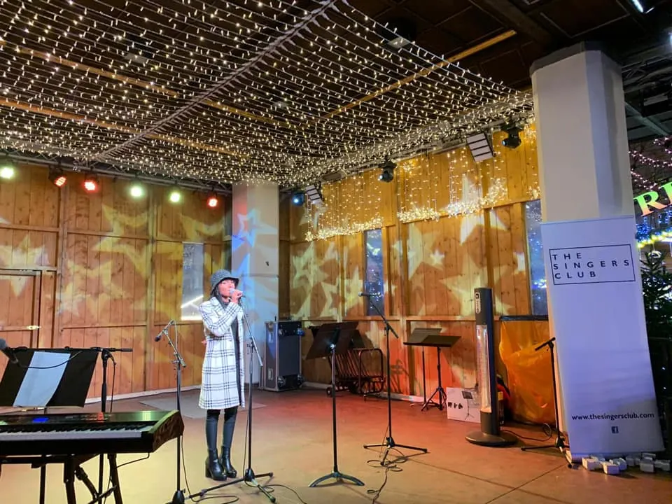 A performer stands at a microphone on stage in a decorated venue with a grand piano, festive lighting overhead, and wooden panelled walls.
