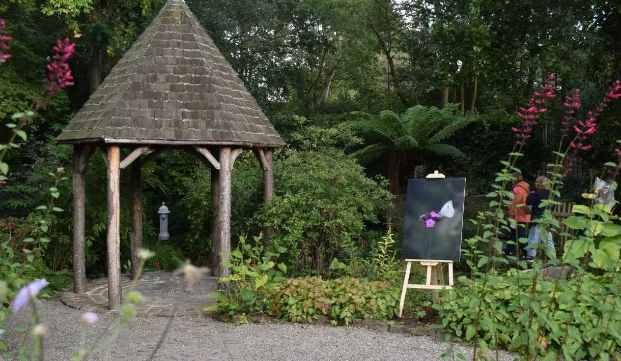 Conical-roofed garden shelter surrounded by flowering plants, shrubs and trees, with an easel displaying artwork nearby.