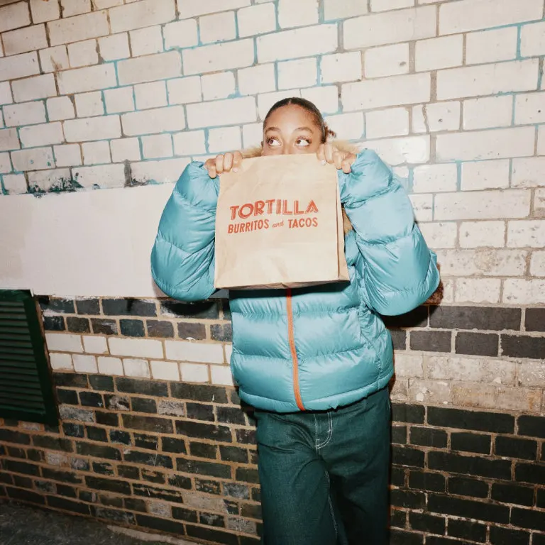 Person in blue puffer jacket holding a branded takeaway bag in front of a brick wall.