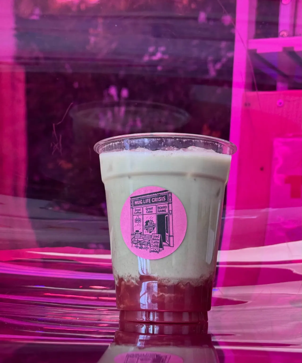 A boba tea drink with dark syrup layer and cream top, labelled with a pink sticker, displayed under bright magenta neon lighting.