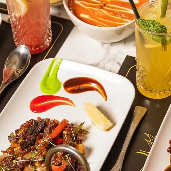 Plated Indian dish with meat and peppers, accompanied by coloured drinks and condiments on a dining table.