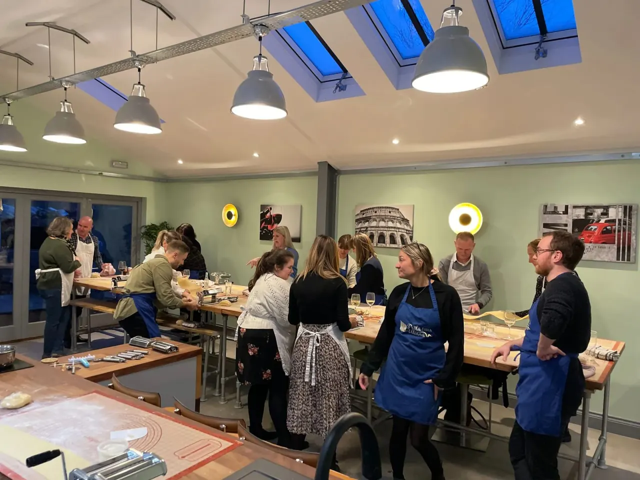 A group of people in blue aprons participate in a pasta-making class at wooden work tables in a bright, modern kitchen studio.