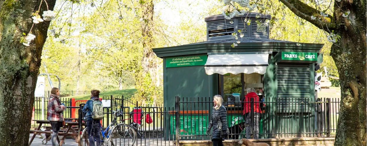 A green café kiosk in a park beneath mature trees, with visitors nearby including someone with a bicycle and children at picnic tables.