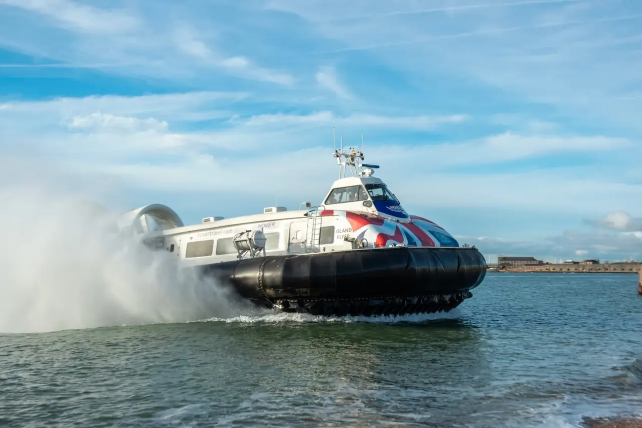A white and blue hovercraft with Union Jack markings approaches a beach in shallow water, spray billowing from its skirt.