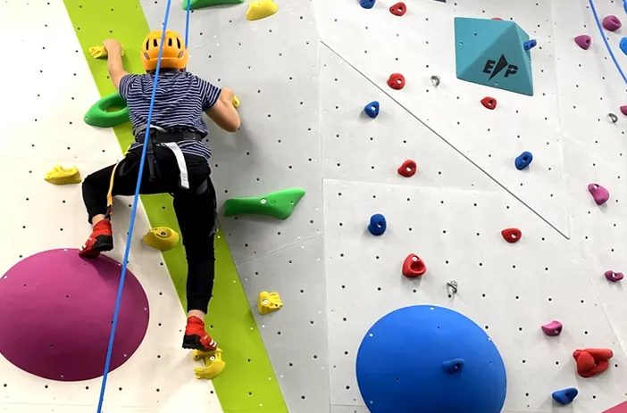 Child rock climbing on an indoor wall with coloured holds, wearing safety harness and helmet.