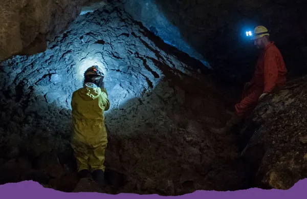 Two visitors in protective gear and headlamps explore an underground cave chamber illuminated by blue lighting.