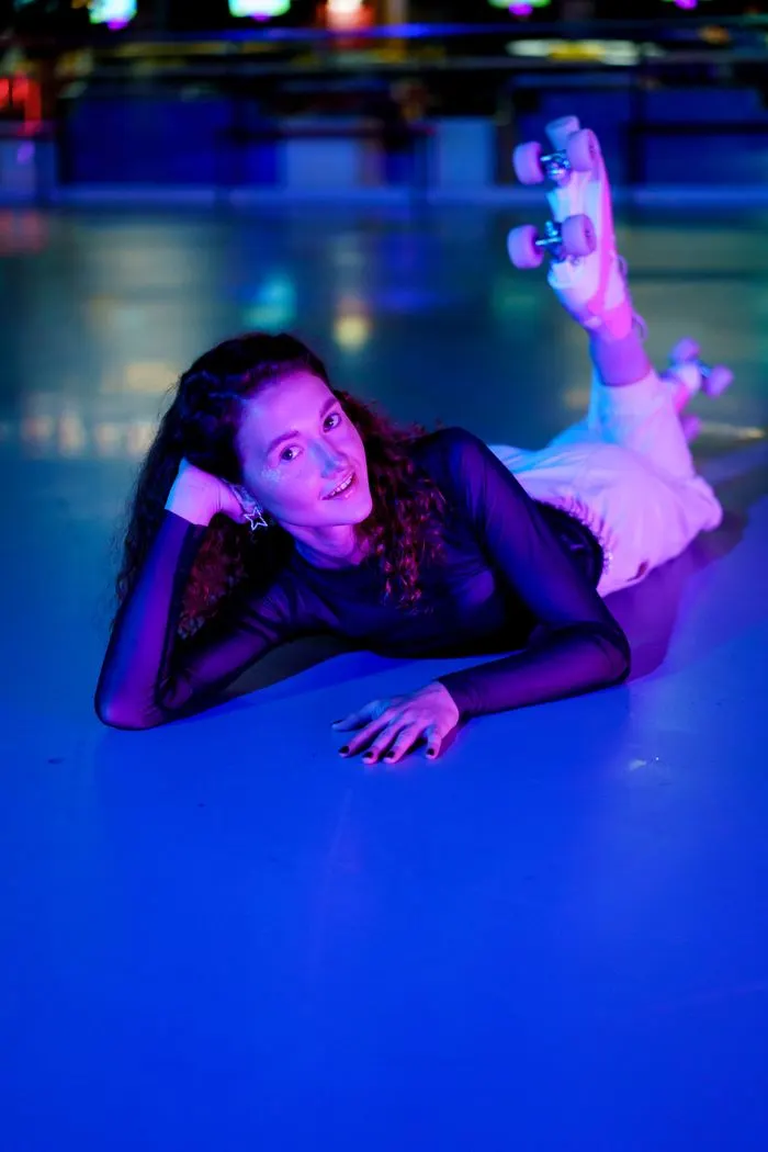 Young person lying on a roller skating rink floor holding a skateboard, lit by purple and blue neon lighting.