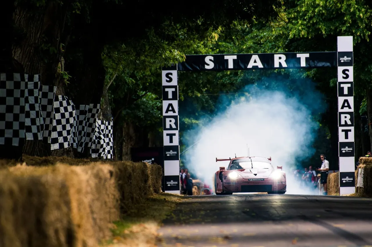 Racing car crosses start line with tyre smoke at motorsport event, flanked by checkered flags and spectators.