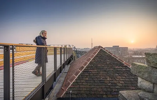 A person stands on a rooftop walkway at sunset, overlooking a city skyline with historic buildings and tiles below.