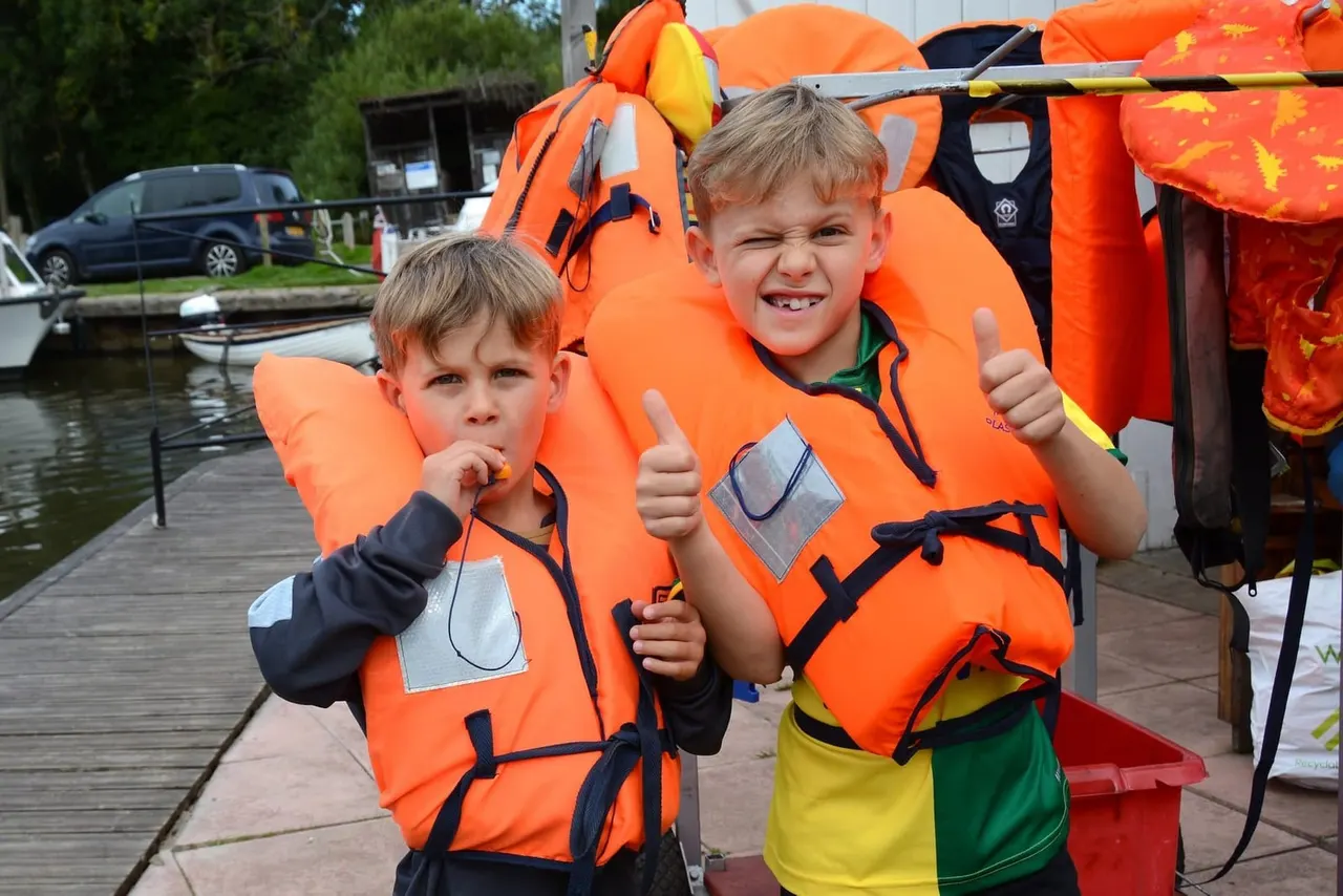 Two young children wearing orange life jackets give thumbs up on a riverside dock.