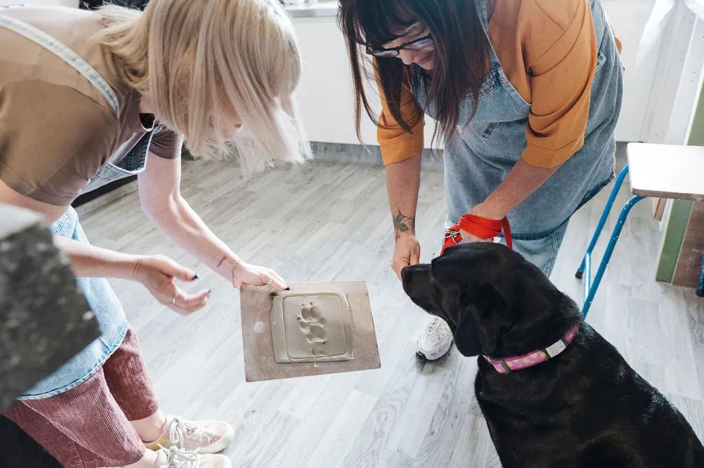 Two people make a paw print impression in clay whilst a black dog watches nearby.