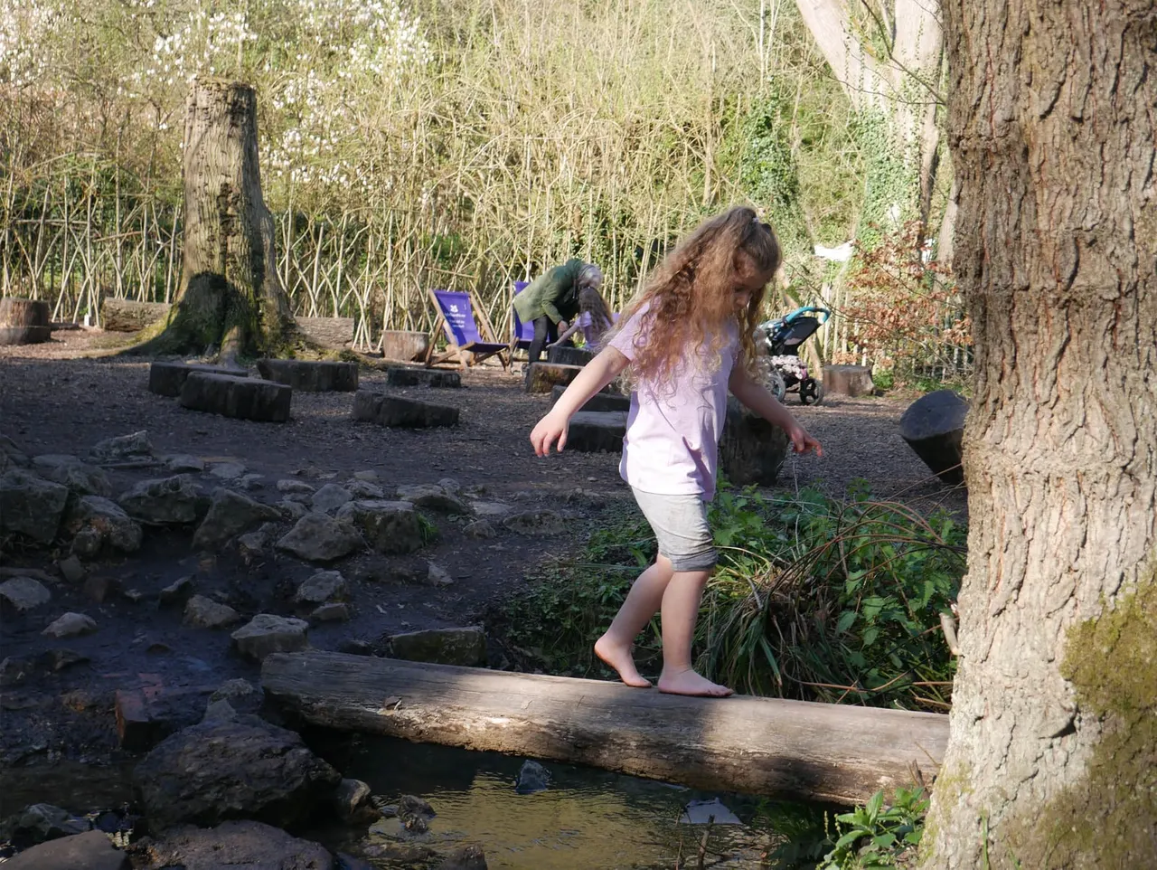 A child balances on a fallen log crossing a stream in a wooded area, with adults relaxing nearby.
