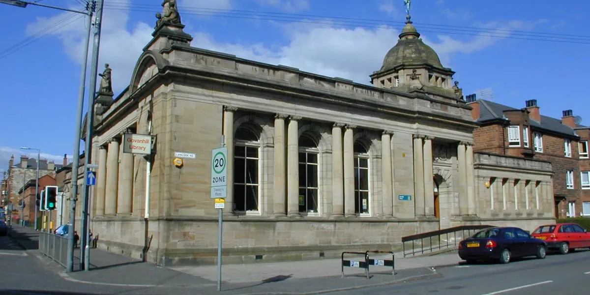 Classical stone library building with columned frontage, domed roof and ornamental finials, situated on a street corner with parked cars and residential tenements nearby.