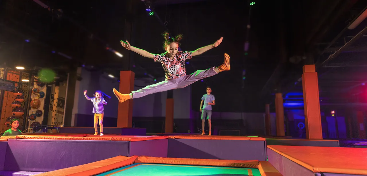 Child performing an aerial jump on a trampoline with arms outstretched whilst other children watch from the padded platform below.