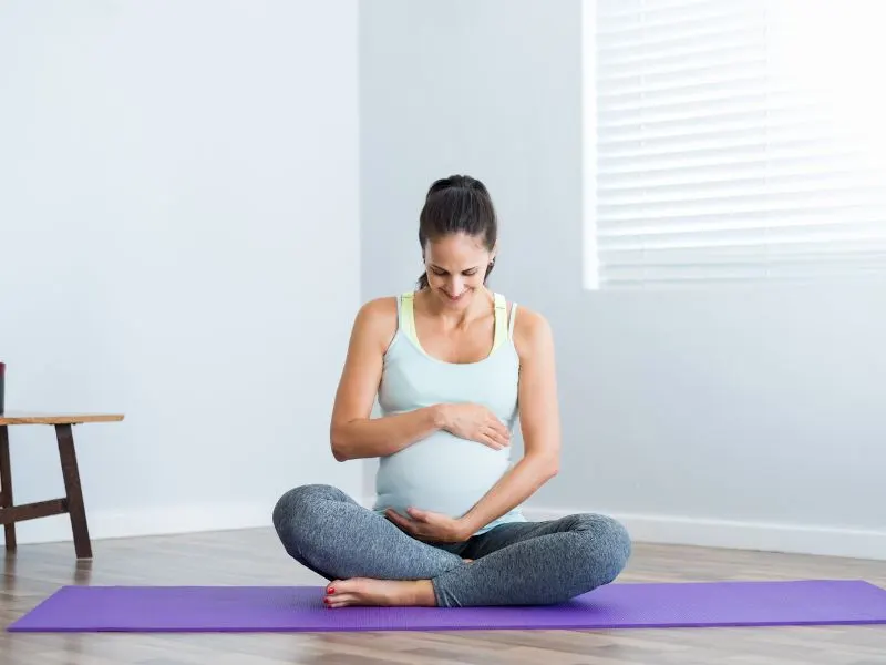 Pregnant woman sitting cross-legged on a purple exercise mat, cradling her belly in a bright, minimalist room.