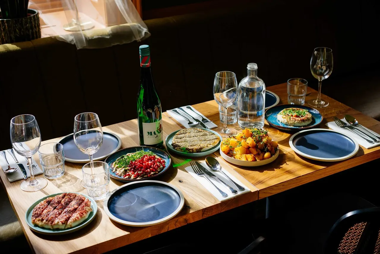 Fine dining table set for four with wine bottle, multiple dishes including flatbreads and roasted vegetables, and place settings with glassware and cutlery.