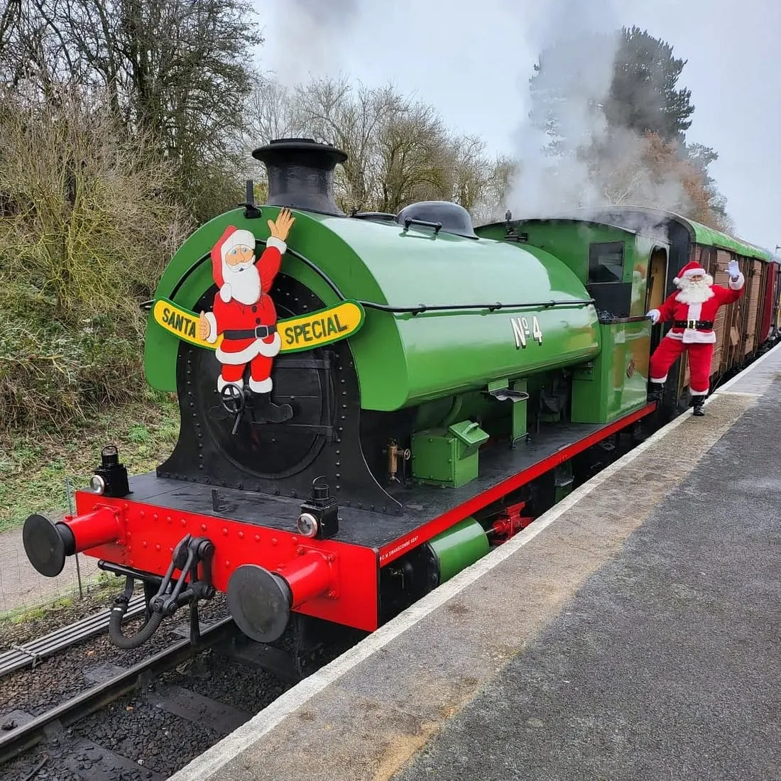 A green and red steam locomotive decorated with Santa designs, emitting smoke, stationed on narrow-gauge railway tracks with a figure in Father Christmas costume visible on the carriage.