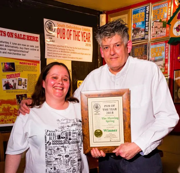 Man and woman holding a "Pub of the Year 2018" award certificate inside a pub decorated with posters and memorabilia.