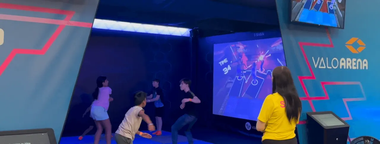 Children playing an interactive video game on a large screen in a brightly lit gaming arena.