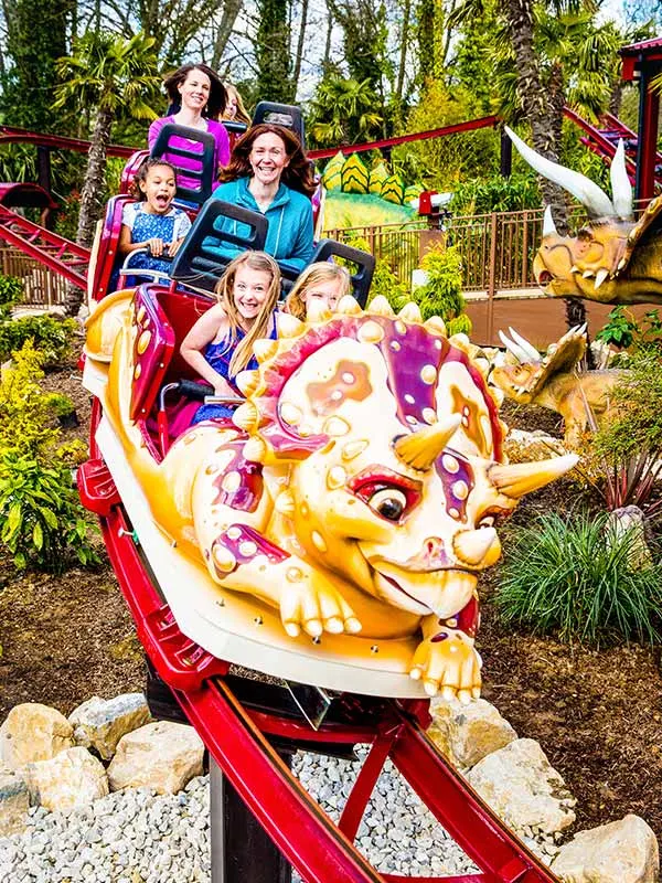 Family of five riding a dinosaur-themed roller coaster with a colourful painted dinosaur head in the foreground.