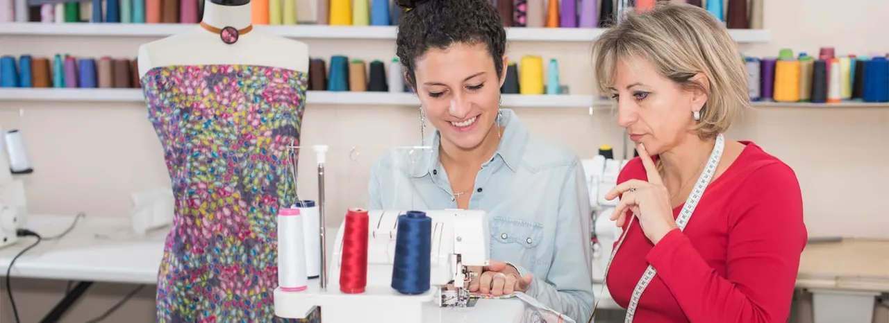 Two women work together at a sewing machine in a craft studio with colourful fabric rolls on shelves behind them.