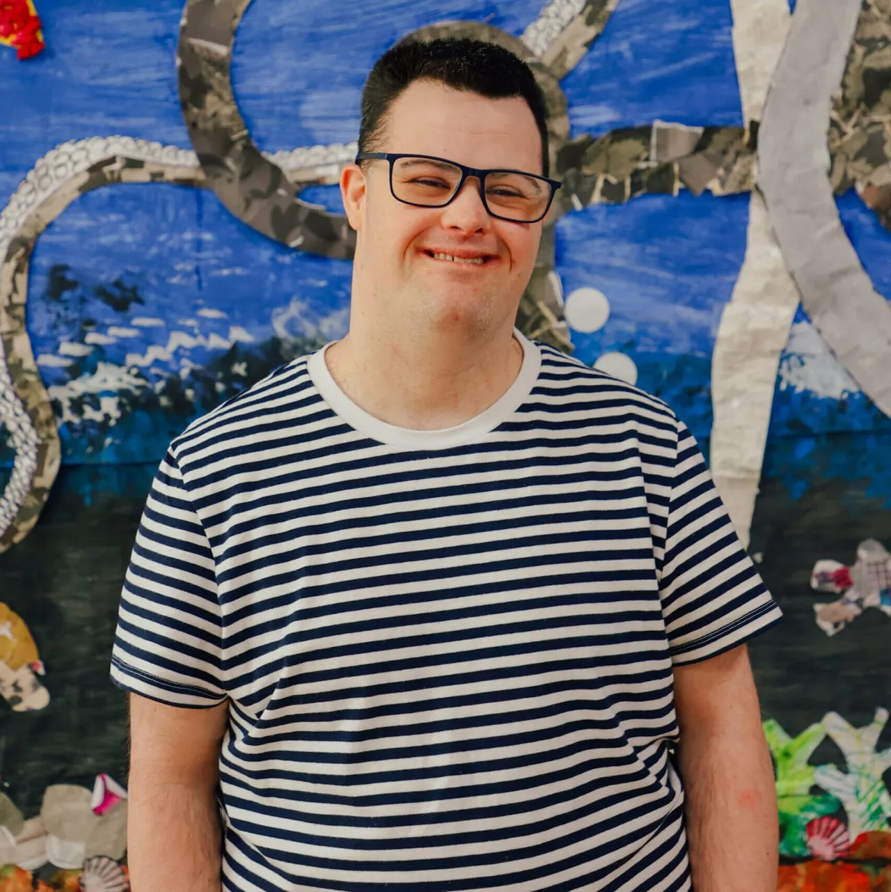 Young man wearing striped shirt and glasses smiling at camera in front of ocean-themed backdrop with coral and sea creatures.