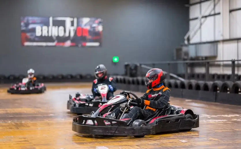 Multiple go-kart drivers racing on an indoor track, wearing helmets and racing suits, with spectator barriers visible.