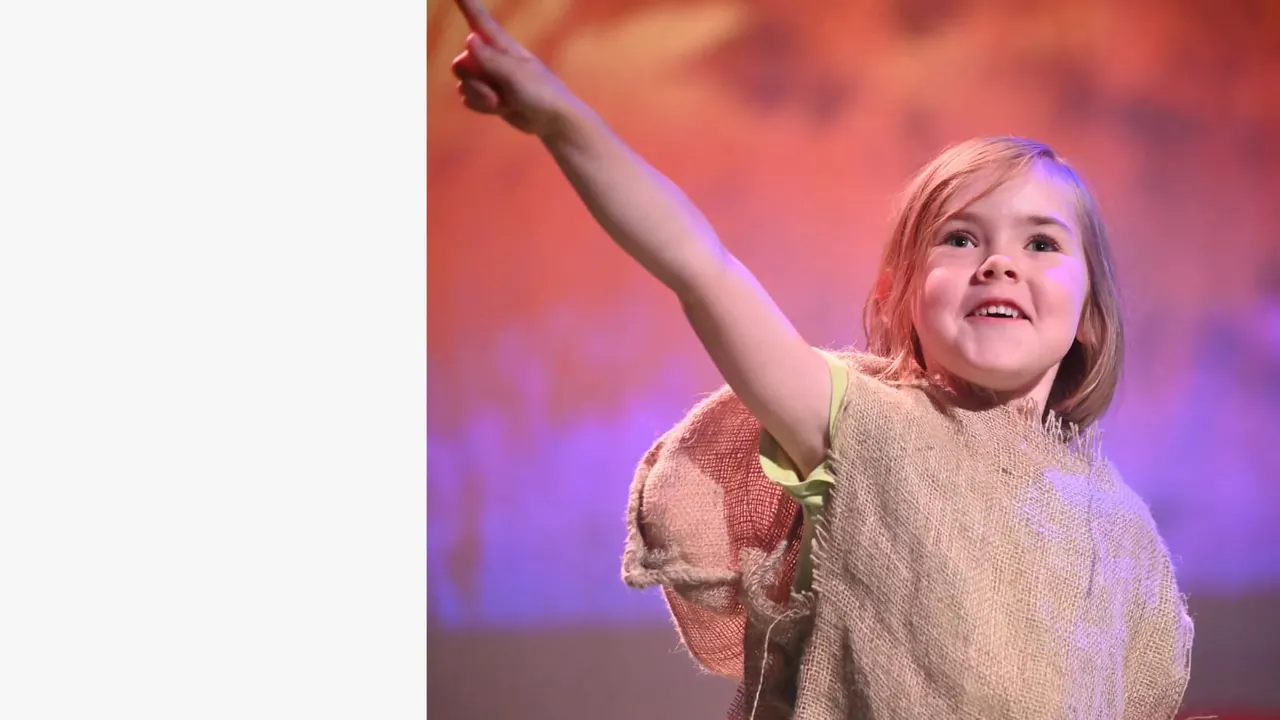 Child on stage pointing upward with a joyful expression under coloured lighting.