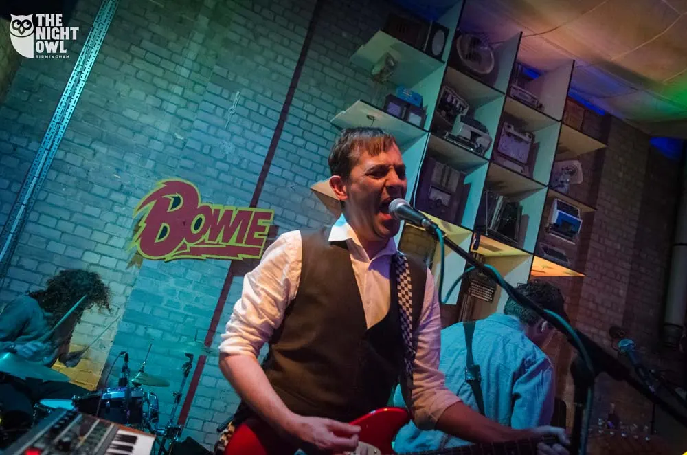 Man singing at microphone on stage in a brick-walled music venue with colourful lighting.