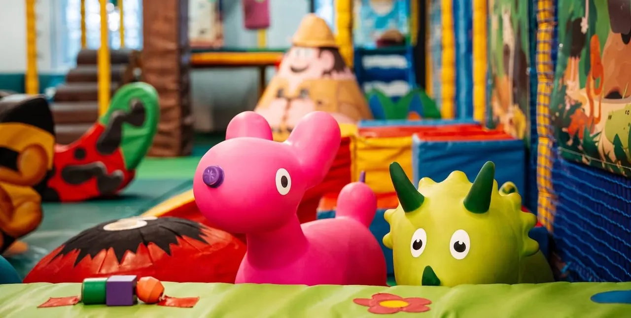 Colourful soft play area with pink and yellow character toys, climbing structures, and bright play equipment for young children.