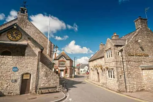 Stone buildings line a quiet village street under blue sky with white clouds.