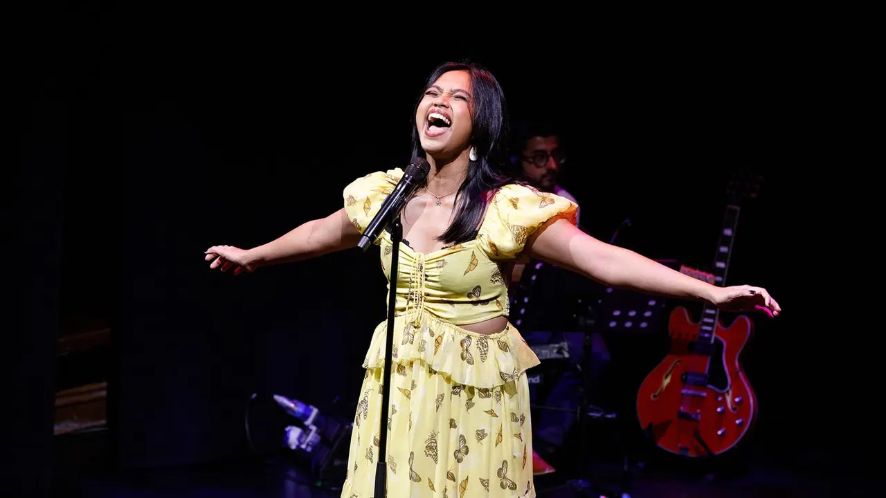 Woman in yellow butterfly-print dress singing at microphone on stage with band and red guitar visible behind her.