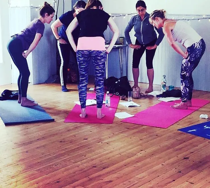 A fitness instructor leads a group exercise class with six adults on coloured mats in a wooden-floored studio.
