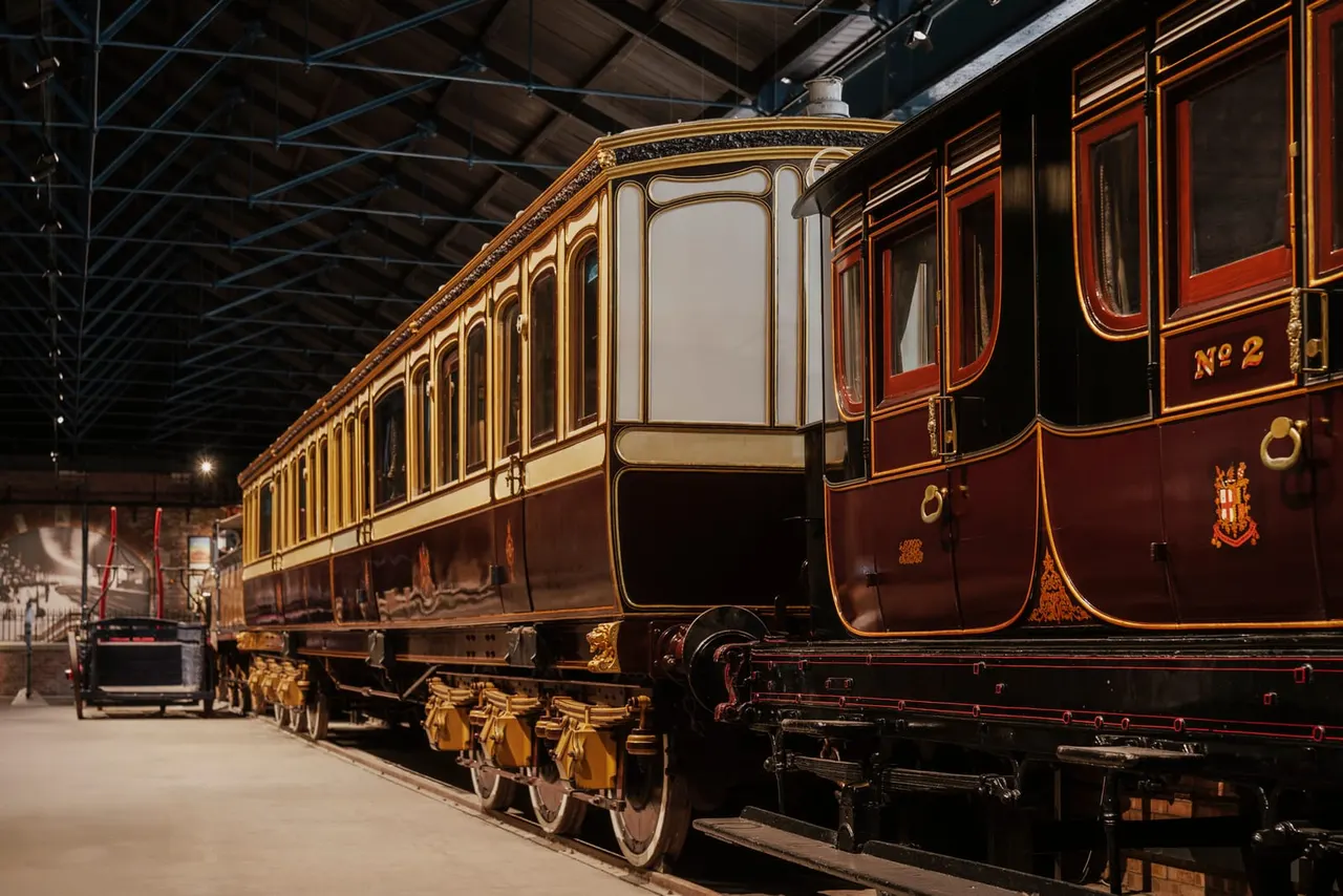 Ornate burgundy and cream railway carriage with gold detailing displayed in a museum shed, showing passenger windows and decorative fixtures.